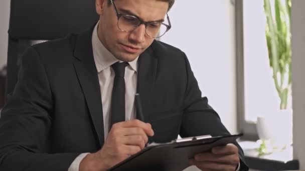 Serious businessman in formal suit and eyeglasses holding clipboard and writing something while sitting in office - Video
