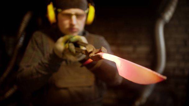 Ένας σιδεράς φτιάχνει ένα μαχαίρι με ένα σφυρί, ψάχνοντας για ατέλειες. - Φωτογραφία, εικόνα