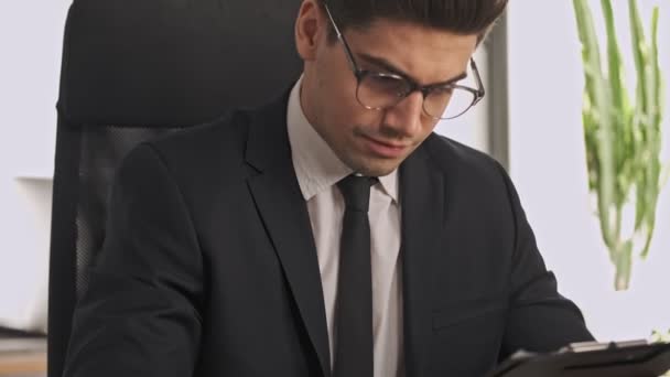 Serious businessman in formal suit and eyeglasses holding clipboard and analyzing something in documents while sitting in office - Video