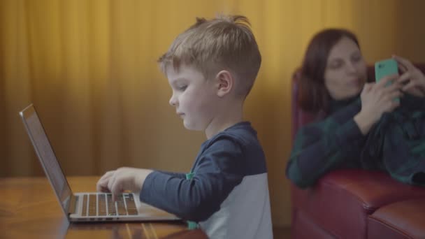 Ξανθιά παιδί προσχολικής ηλικίας δακτυλογράφηση σε φορητό υπολογιστή και χαμογελώντας κοιτάζοντας οθόνη υπολογιστή. Μητέρα με κινητό τηλέφωνο που βρίσκεται στον καναπέ στο παρασκήνιο. Παιδί μαθαίνει online στο σπίτι κάθεται στο τραπέζι.  - Πλάνα, βίντεο