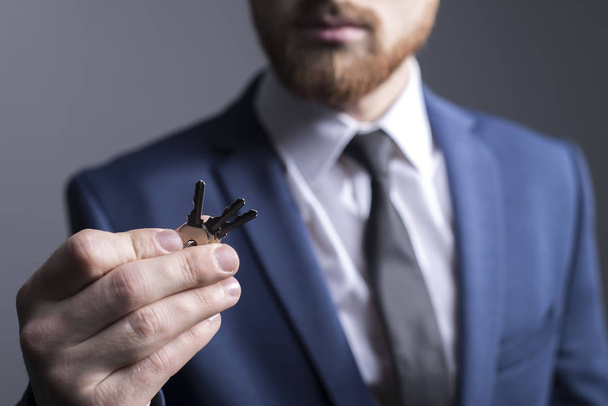 close-up portrait of a young bearded guy of twenty-five years old, in a business suit, holding keys in his hand. On a gray background. concept. purchase of a new apartment, keys to the safe, in focus a hand holding a key, a business suit and tie. - Photo, image