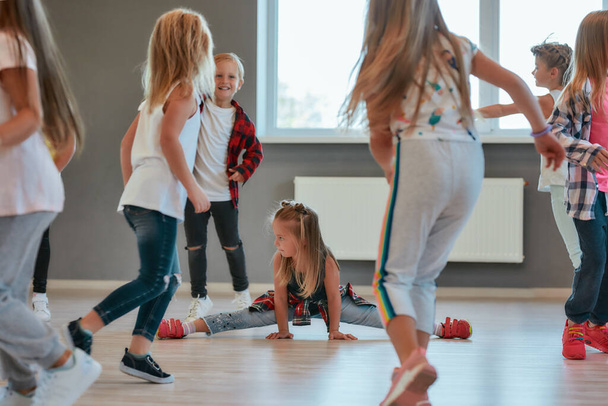 Gymnastic exercises. Little cute girl sitting on a twine while having a choreography class. Group of positive and active children learning a modern dance in the dance school - Foto, immagini
