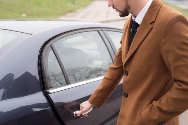 Portrait of a young guy in business clothes, a businessman of thirty years opens the car door to exit. In an interesting authoring. A bearded businessman is driving in a car to work in an office. - Foto, afbeelding