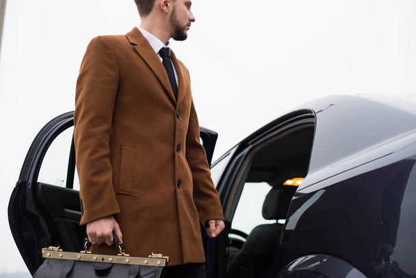 Portrait of a young bearded guy thirty years old. A businessman in a business suit, with a tie, stands against the backdrop of a car. Business concept business style, office style. Business man in a taxi car standing - Photo, image