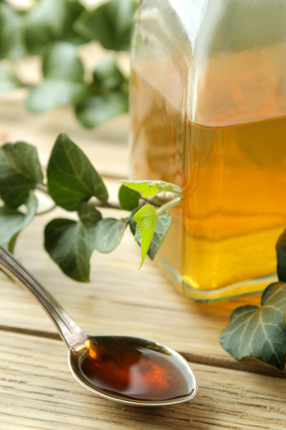 Φύλλα κισσού και σιρόπι σε ένα μπουκάλι σε ένα φυσικό ξύλινο τραπέζι. Παραγωγή αντιβηχικού φαρμάκου με εκχύλισμα κισσού. Φαρμακευτική βιομηχανία - Φωτογραφία, εικόνα