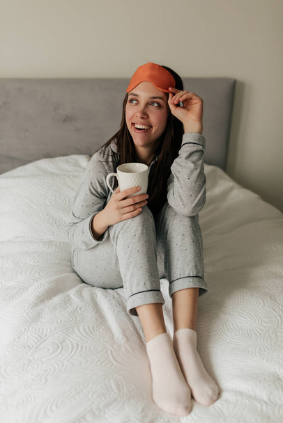 Opgewonden charmant meisje in pyjama en slaapmasker wakker, zittend op het slechte met de ochtend kopje koffie. Fijne dag thuis. - Foto, afbeelding