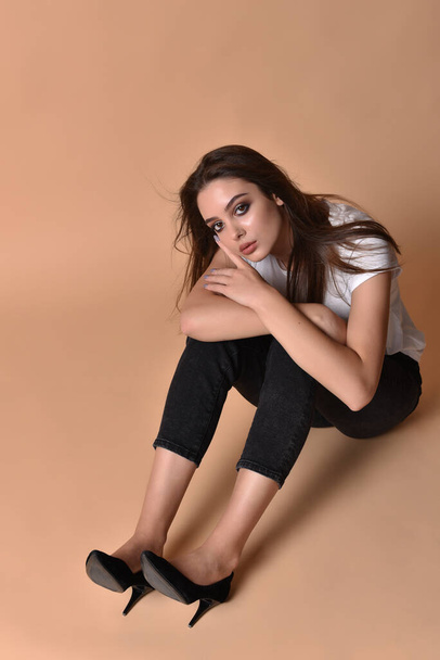 Sexy brunette woman posing. Girl with professional make-up.Pretty woman posing with hands in the frame in a white T-shirt and bright professional makeup. The girl is sitting on the studio floor - Φωτογραφία, εικόνα