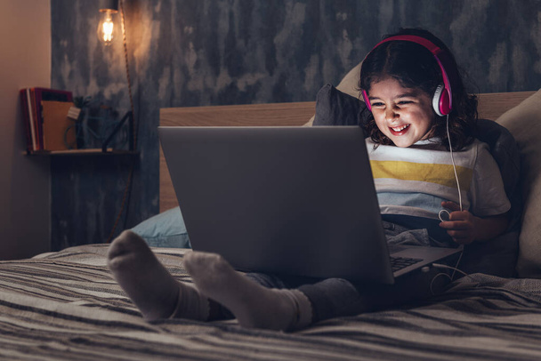 little girl sitting on the bed between cushions laughing while playing or watching a movie on a computer in the darkness, has a pink headset, child and technology concept - Foto, immagini