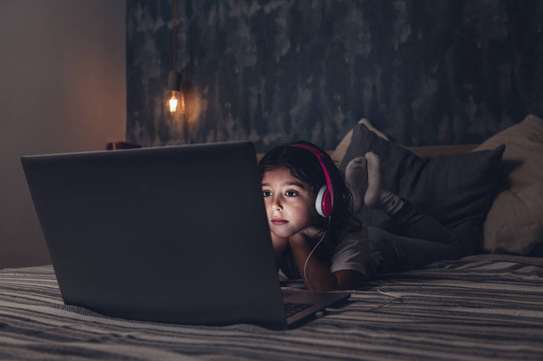 dziewczynka leżąca w łóżku skupiona podczas oglądania filmu na laptopie w ciemności, ma różowy zestaw słuchawkowy, koncepcję dziecka i technologii, kopiuj miejsce na tekst - Zdjęcie, obraz