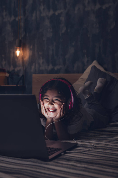 vertical photo of a happy little girl lying in bed smiling while watching a movie on a computer in the darkness, has a pink headset, child and technology concept, copy space for text - Foto, Imagem