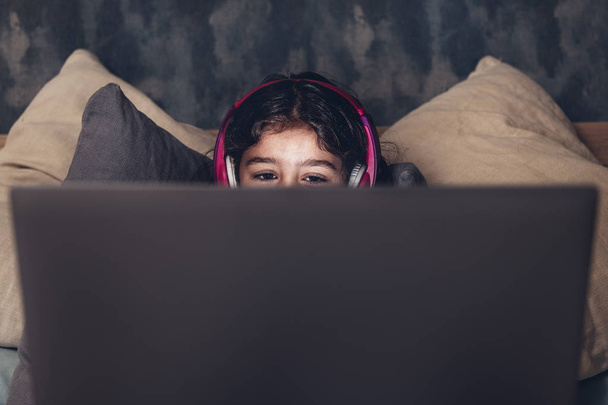detail of the eyes of a little girl on the bed focused while playing or watching a movie on a computer in the darkness, has a pink headset, child and technology concept, copy space for text - Foto, Imagem