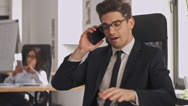 Cheerful businessman in formal suit and eyeglasses talking by smartphone while sitting by the table in office - Imágenes, Vídeo