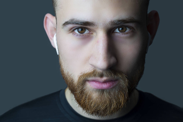 Close-up portrait of a young bearded guy of twenty-five years old, looking at the camera. In wireless white headphones. Sporty style. Athletes headphone promotional photo - Fotoğraf, Görsel