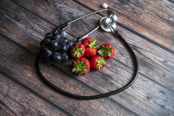 A closeup shot of strawberries and blueberries surrounded by a stethoscope on a wooden table - Zdjęcie, obraz
