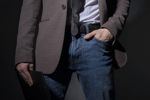 Close-up of a guy in jeans and a formal suit, holds a folder in his hand. Business style. On a dark background. Art light. Advertising photo of mens clothing. Male fashion look - Valokuva, kuva