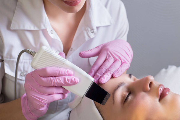closeup, professional beautician, using an apparatus for ultrasonic face cleaning, cleans the skin of a girl patient, in a beauty salon. Master in cosmetology portrait. Professional and medical facial skin care, when performing procedures that improv - Photo, Image