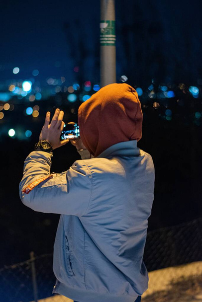 A man wearing a hoodie taking the picture of the night view of a city - Φωτογραφία, εικόνα