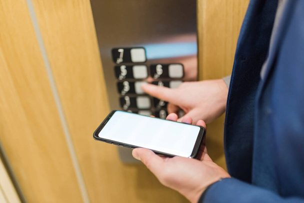 Man hands holding blank screen mobile phone while using elevator control panel. - Фото, изображение