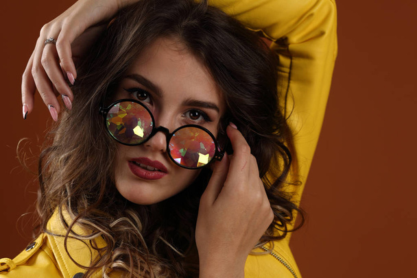Studio portrait of a beautiful girl in a yellow jacket and glasses with colored glasses on a brown background - Φωτογραφία, εικόνα