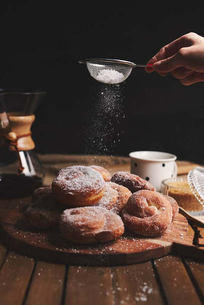 A vertical shot of delicious snake doughnuts sprinkled with powdered sugar and chemex coffee on a wooden table with a black blurred background - Fotografie, Obrázek