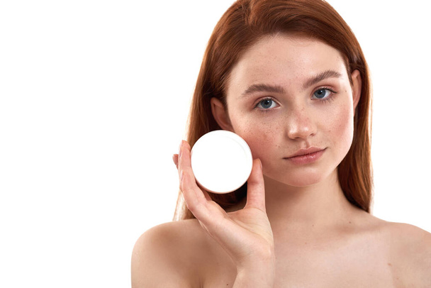 My skincare routine. Young attractive red-haired girl with freckles demonstrating cosmetic product and looking at camera while standing in studio against white background - Photo, Image