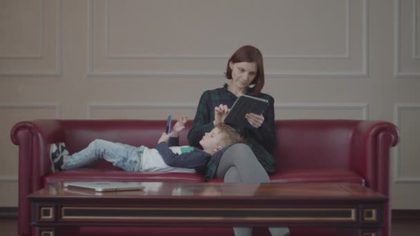 Young 30s mother with tablet computer and boy laying on her leg using gadget on couch. Family watching online content on different devices at home.  - Materiał filmowy, wideo