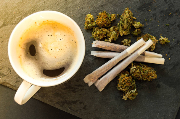 Delicious cup of coffee, a pile of high quality marijuana buds with some cigars of weed ready to smoke. Top view with background of black stone. - Photo, Image