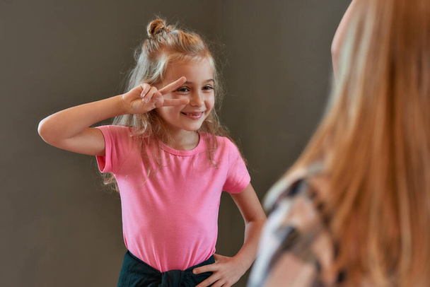 Portrait of pretty little girl in the pink t-shirt showing a peace gesture and smiling while standing against grey wall and having choreography class in the dance studio. - Foto, afbeelding