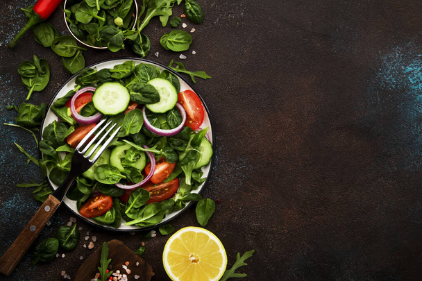 Healthy food background. Ingredients for cooking salad. Cherry tomatoes, spinach, arugula, cucumbers, spices and oil with plate on brown background. Top view, copy space - Foto, afbeelding