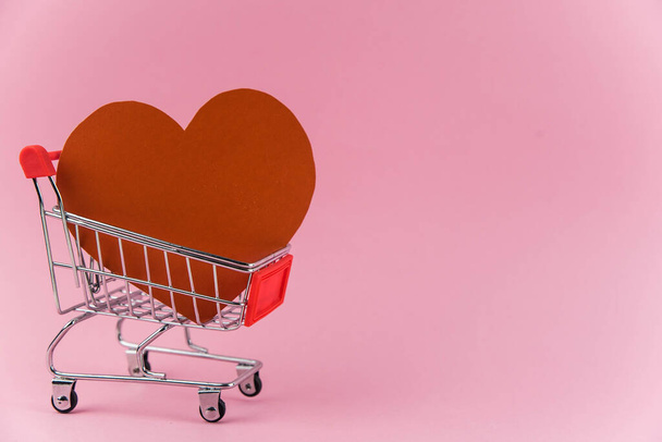 Red heart in a shopping cart on trendy pink background with copy space for text. Concept of holiday Valentine`s day shopping, sale and waste of money for love  - Photo, Image