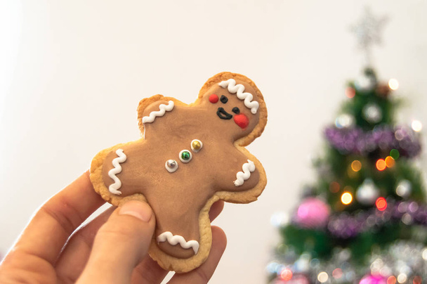 Close-up de uma mão segurando um biscoito de Natal caseiro com árvore de Natal borrada com luzes no fundo. Casa com decoração de Natal
. - Foto, Imagem