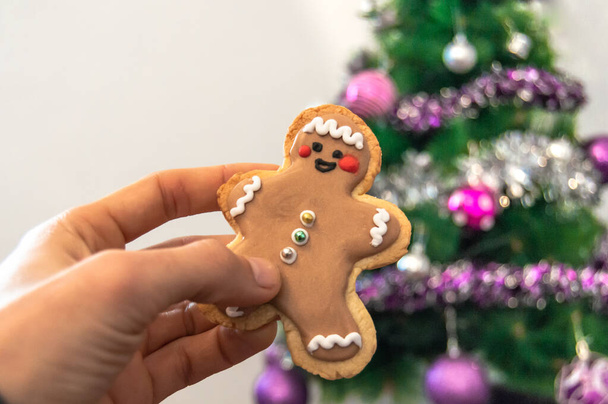 背景にライトが付いているぼやけたクリスマスツリーと自家製のクリスマスクッキーを持っている手のクローズアップ。クリスマスの装飾付きの家. - 写真・画像