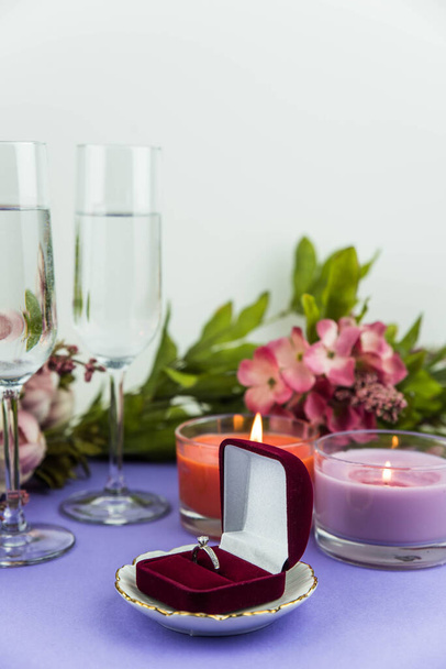 Huwelijk aanzoek en liefde concept voor Valentijnsdag, doos met bruiloft of verlovingsring met briljant. Glas champagne, romantisch diner, pioenroos bloemen en kaarsen op trendy paarse achtergrond - Foto, afbeelding
