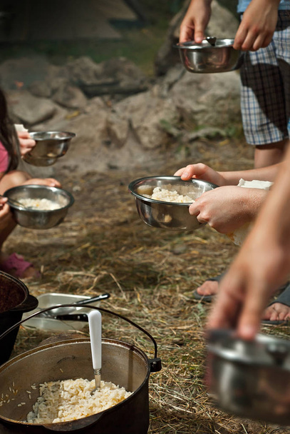 Kids eating on a camping trip. The food in the cauldron is cooked on fire. Girl and boy holding a mask. Children throw food together in plates. - 写真・画像