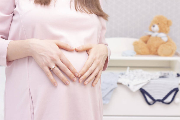 pregnant woman in a dress holds her hands on her stomach on the background of things for the newborn and a toy bear. Pregnancy, motherhood, preparation and expectation concept. Close-up indoors. - Foto, imagen