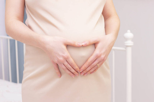 A pregnant woman in a dress holds her hands on her stomach in the shape of a heart. The concept of pregnancy, motherhood, preparation and expectation. Close-up indoors. - Photo, Image