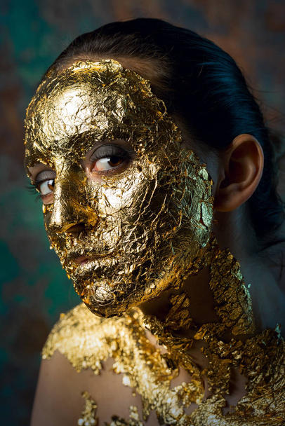 Girl with a mask on her face made of gold leaf. Gloomy studio portrait of a brunette on an abstract background. - Фото, зображення
