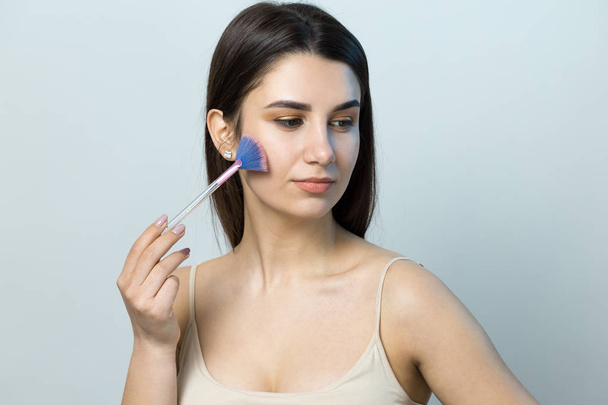 Close-up of a young girl in a light top on a white background making a facial make-up. A pretty woman holds a cosmetic brush near her face and smiles. - Foto, Imagem