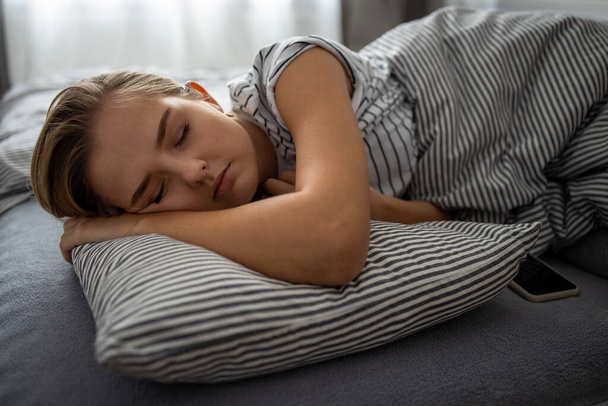 Mooie, jonge vrouw slaapt in haar bed met haar mobiele telefoon dicht bij haar. Smartphone in bed Mobiel / smartphone Verslavingsconcept. - Foto, afbeelding