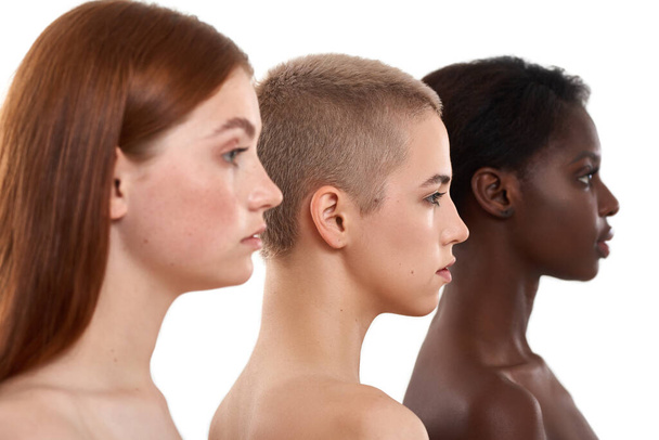 Different skin tones. Side view of three beautiful multicultural young women.Two caucasian and african half naked women posing in studio over white background. Natural beauty - Фото, изображение