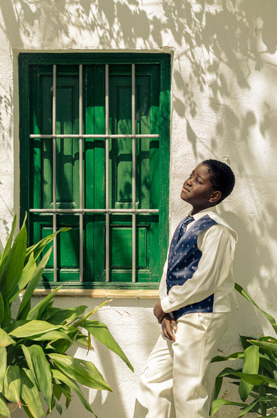 Black boy with white shirt and blue tie next to a green window - Фото, изображение