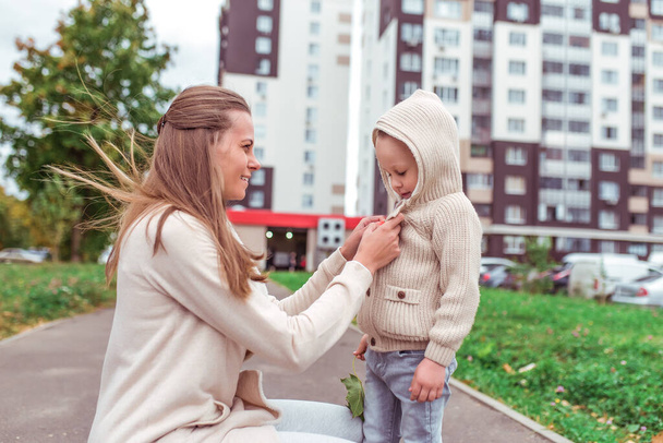 Woman mother, fastens sweater to son of boy, autumn day, street city, strong wind, child jumper with hood. Care care upbringing, emotions of tenderness of parenting in front of beloved child. - Fotoğraf, Görsel