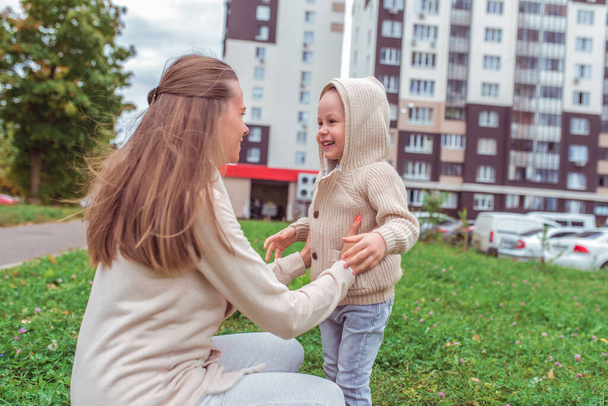Happy family, woman mom, plays with her little son boy, summer in city on street. Emotions of fun, joys of laughter and smile. Casual warm clothes with a hood. Long hair, child care and parenting. - Foto, Bild