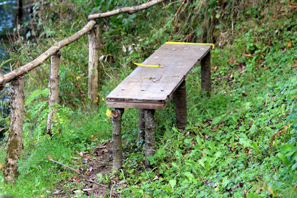 Improvised homemade bench made from wooden board supported with cut wooden sticks next to small fence at local river bank surrounded with uncut grass and small plants - Photo, Image