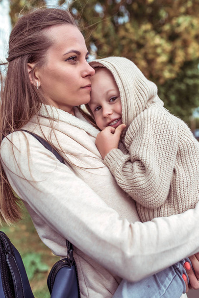 Young family, woman mom, holds in her arms child, little son boy, in fall. Walk in city. Emotions of comfort pleasure, smile. Casual warm clothes with hood. Caring for child and parenting. - Valokuva, kuva