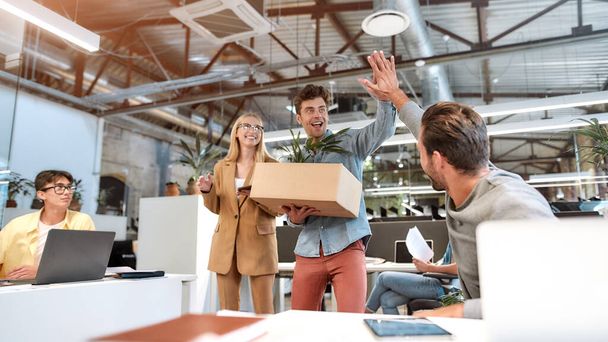 Welcome on board. Young man in casual wear holding box with things and giving high five to his new coworker while standing in the modern office with other colleagues - Foto, Imagen
