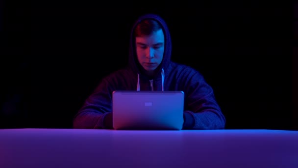 Young man in a hood with a laptop. Hacker makes a hack through a laptop. Blue and red light falls on a man on a black background. - Záběry, video