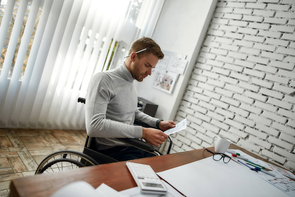 Young and handsome male engineer or architect in a wheelchair looking at blueprint while sitting at his working place in the modern office - Fotografie, Obrázek