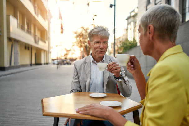 Early morning. Happy senior couple enjoying coffee while sitting at cafe outdoors - 写真・画像