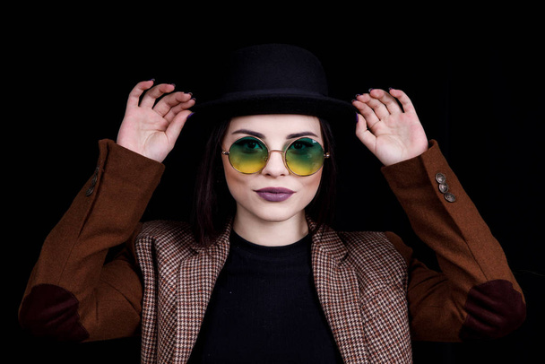 Красивая девушка в шляпе и круглых очках позирует в студии на черном фоне
 - Фото, изображение